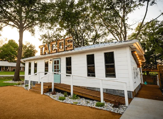 The Doc's Taco Shop building in Liberty City, Texas - white clapboard with a rustic TACOS marquee sign on the roof, a mint green door and string lights in the trees