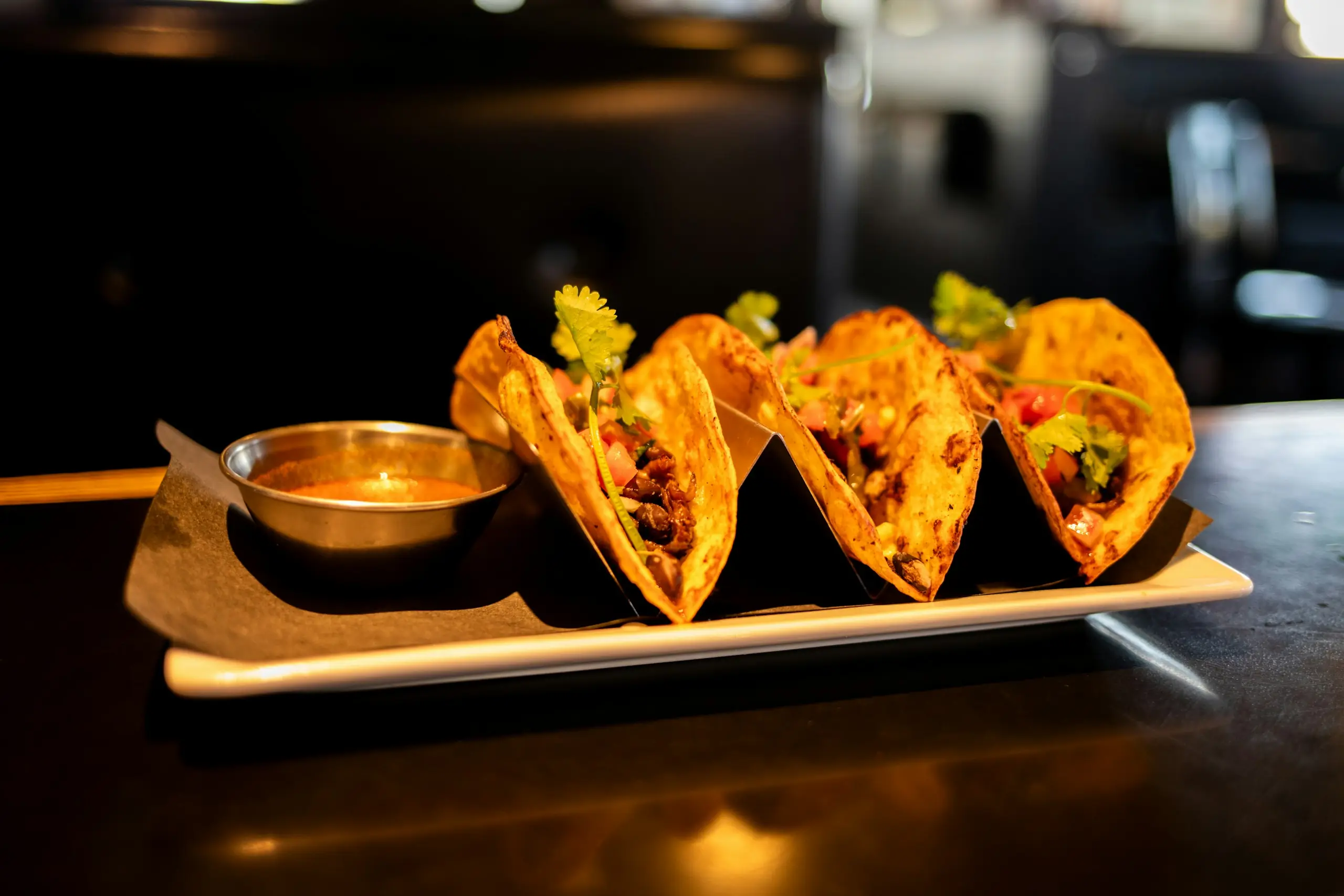 Three street tacos with cilantro and pico de gallo on a plate with salsa, lit warmly in a cantina