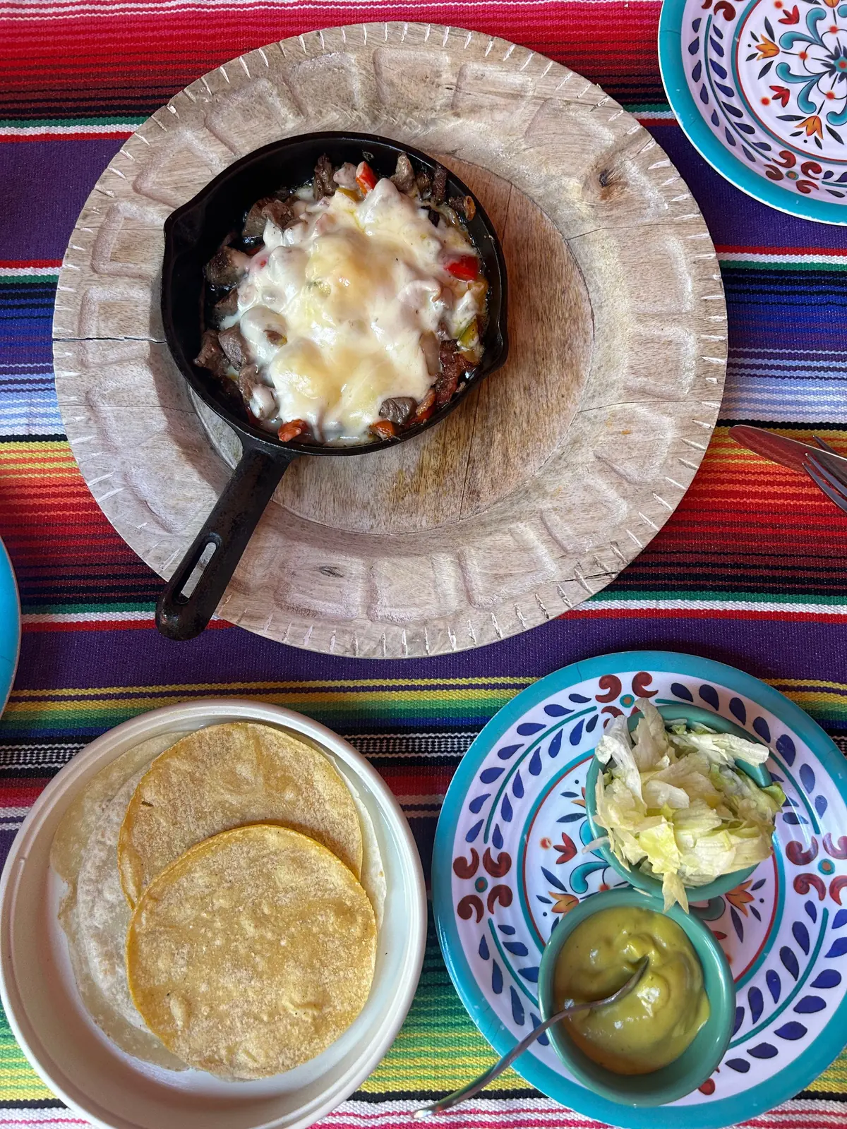 An overhead spread of Mexican food on a striped serape cloth — a cast-iron skillet, warm corn tortillas, guacamole and salad on talavera plates