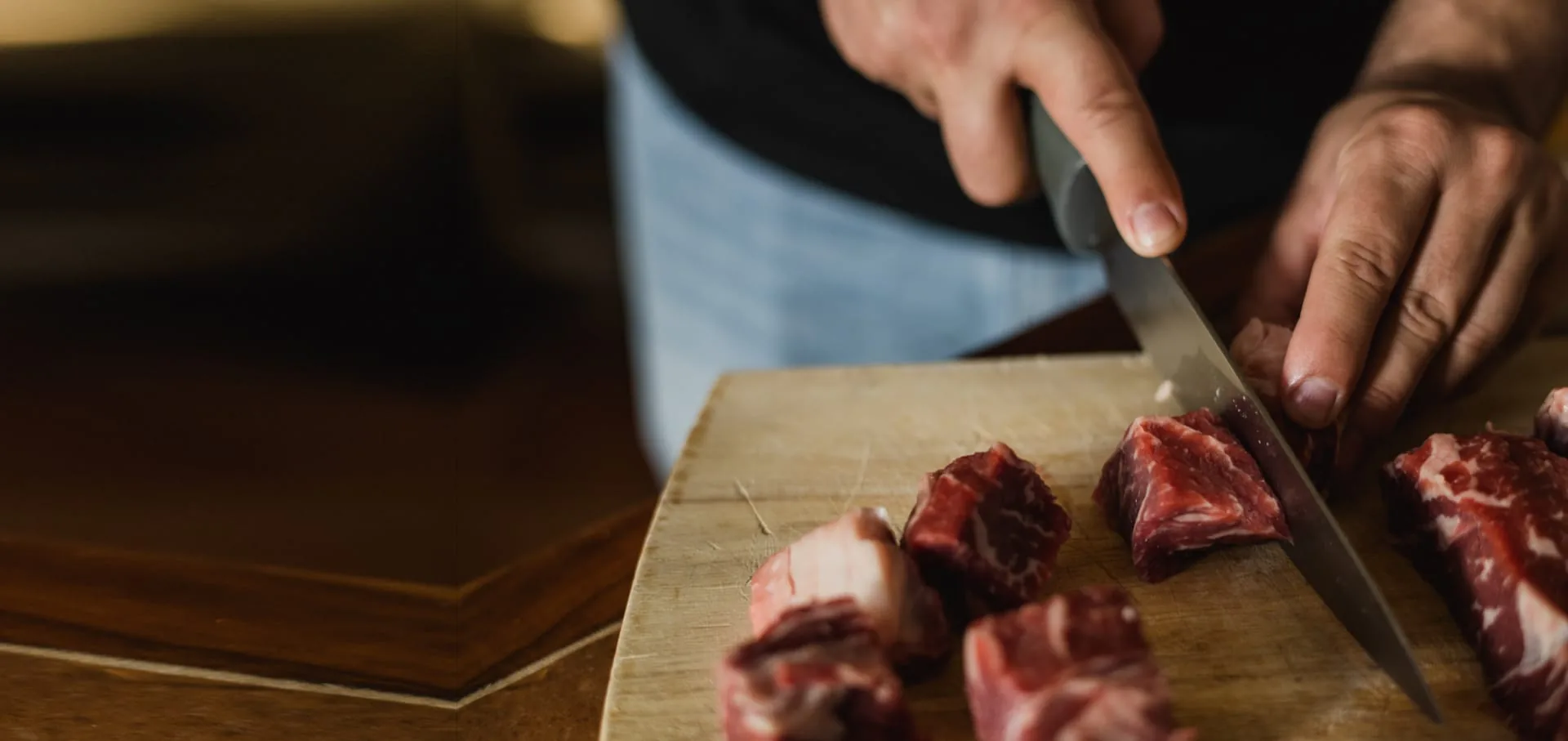 Hands slicing beef into cubes on a wooden board in the kitchen