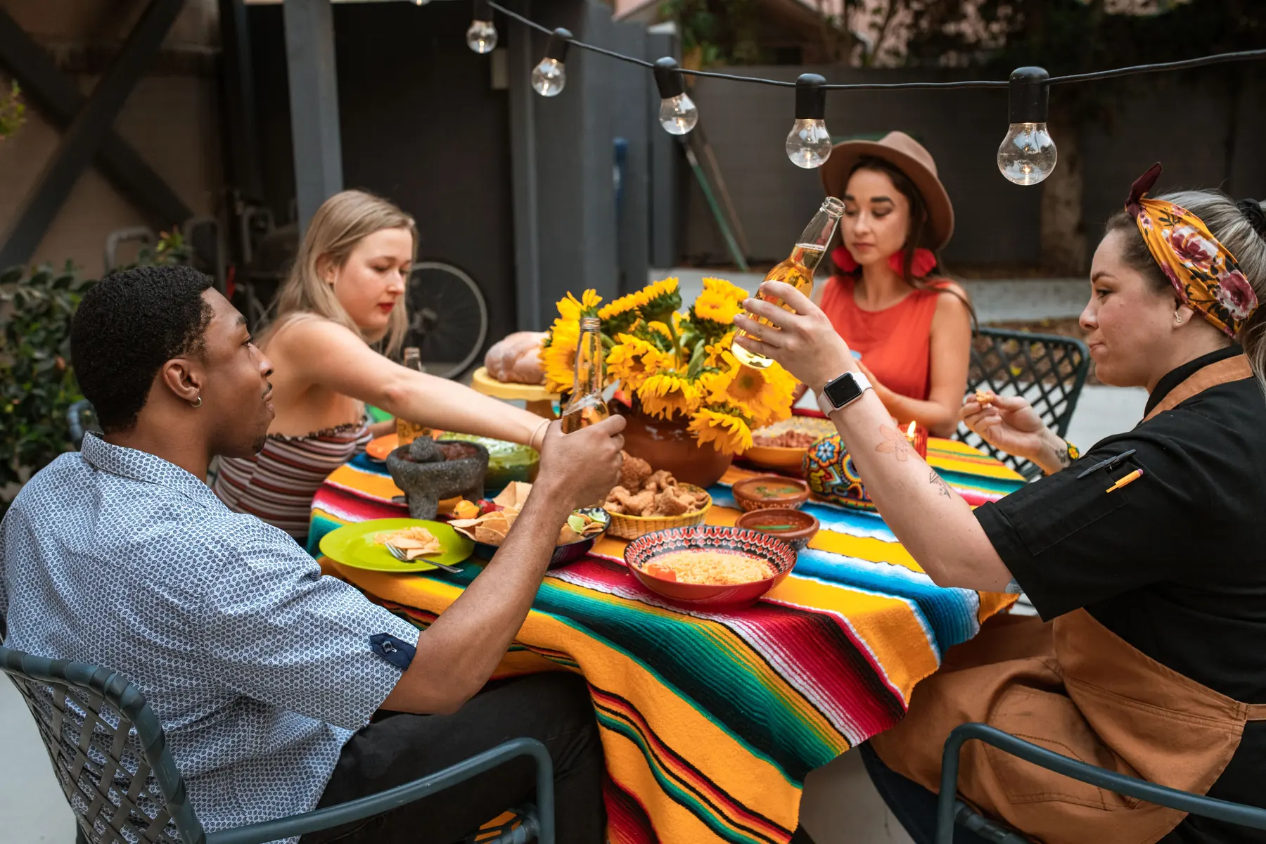 Friends sharing a Mexican meal at an outdoor table under string lights, with salsa, chips and sunflowers