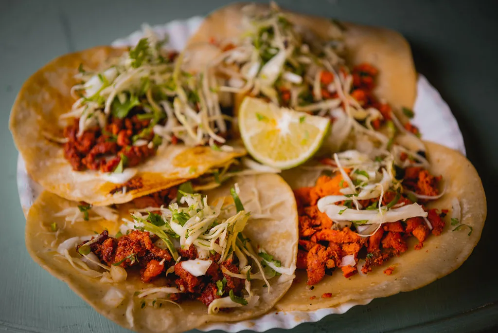 A paper plate of Doc's Taco Shop al pastor street tacos on corn tortillas with shredded cabbage, cilantro, onion and a lime wedge