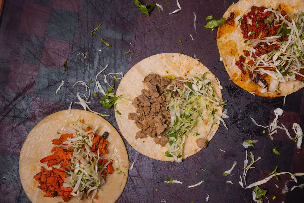 Three open street tacos topped with shredded cabbage and cilantro, filled with al pastor, bistek and pollo